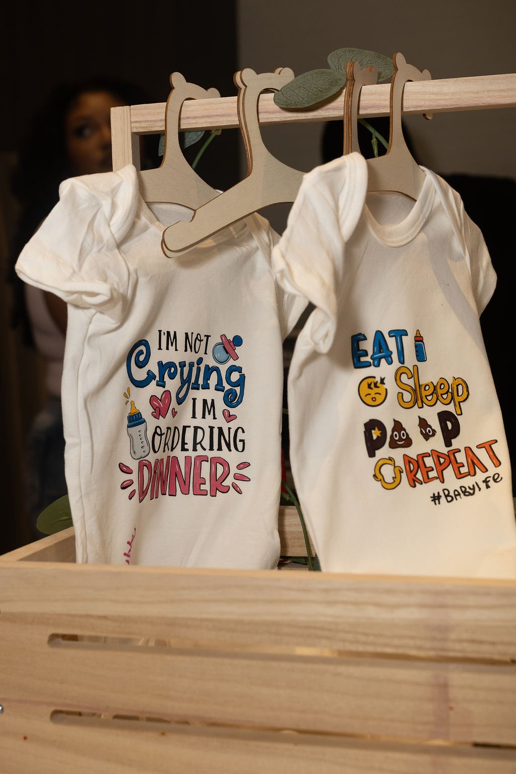Two finished onesies hand-decorated by guests at the DIY design-a-bodysuit station, reading 'I'm not crying I'm ordering dinner' and 'Eat sleep poop repeat'