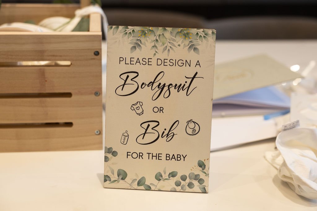 Sign inviting guests to design a bodysuit or bib for the baby at the DIY décor station