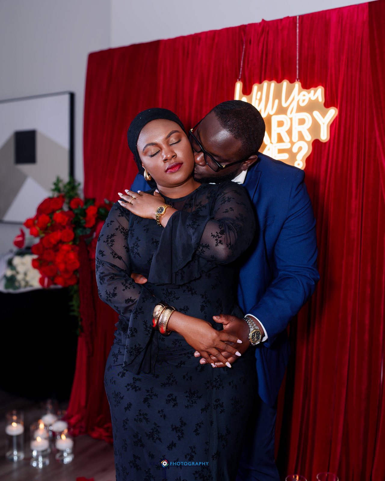 The couple sharing a warm embrace in front of the red rose backdrop