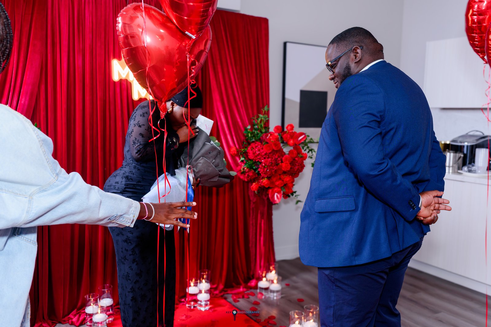 The groom presenting a rose bouquet to the bride beside red heart balloons during the surprise