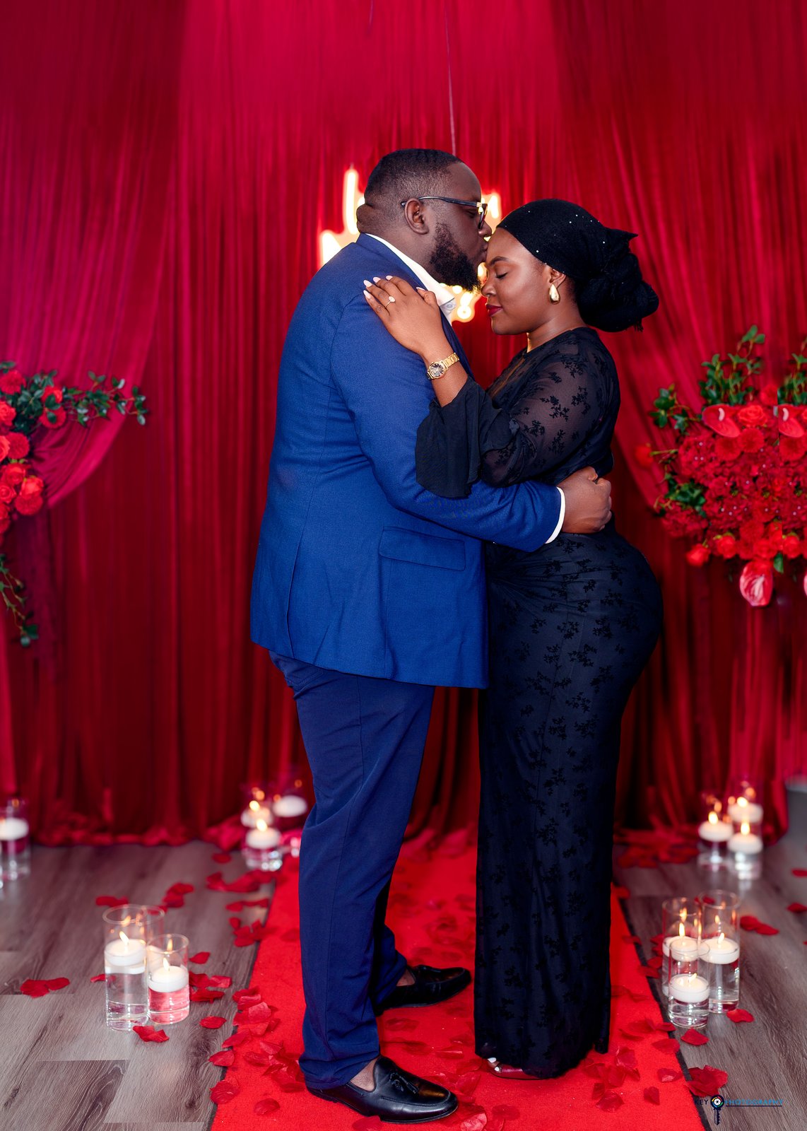 The couple embracing closely in front of the red rose backdrop