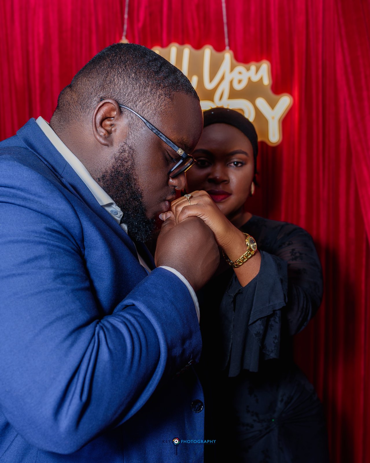 The groom tenderly kissing the bride's hand beneath the Will You Marry Me neon sign