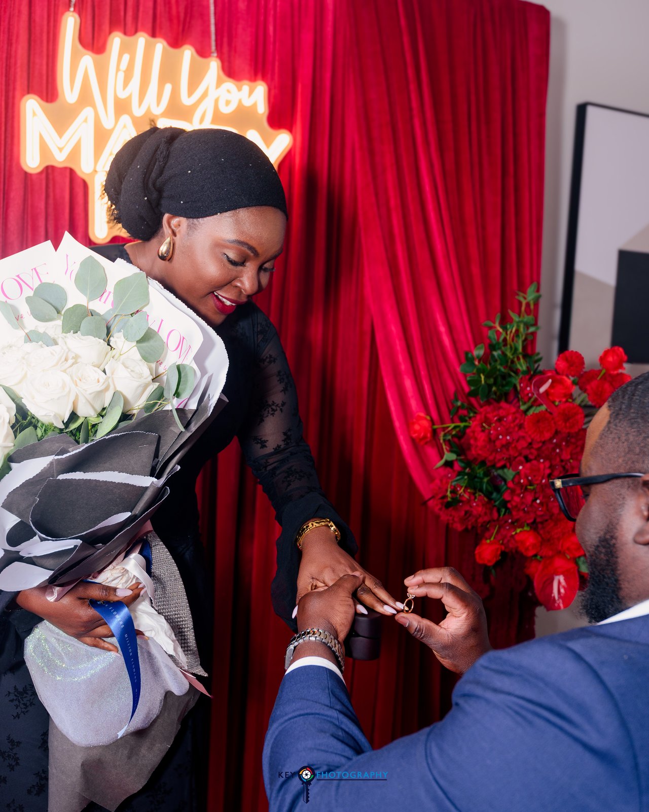 The groom placing the ring on the bride's finger as she holds her white rose bouquet, red curtain backdrop behind them