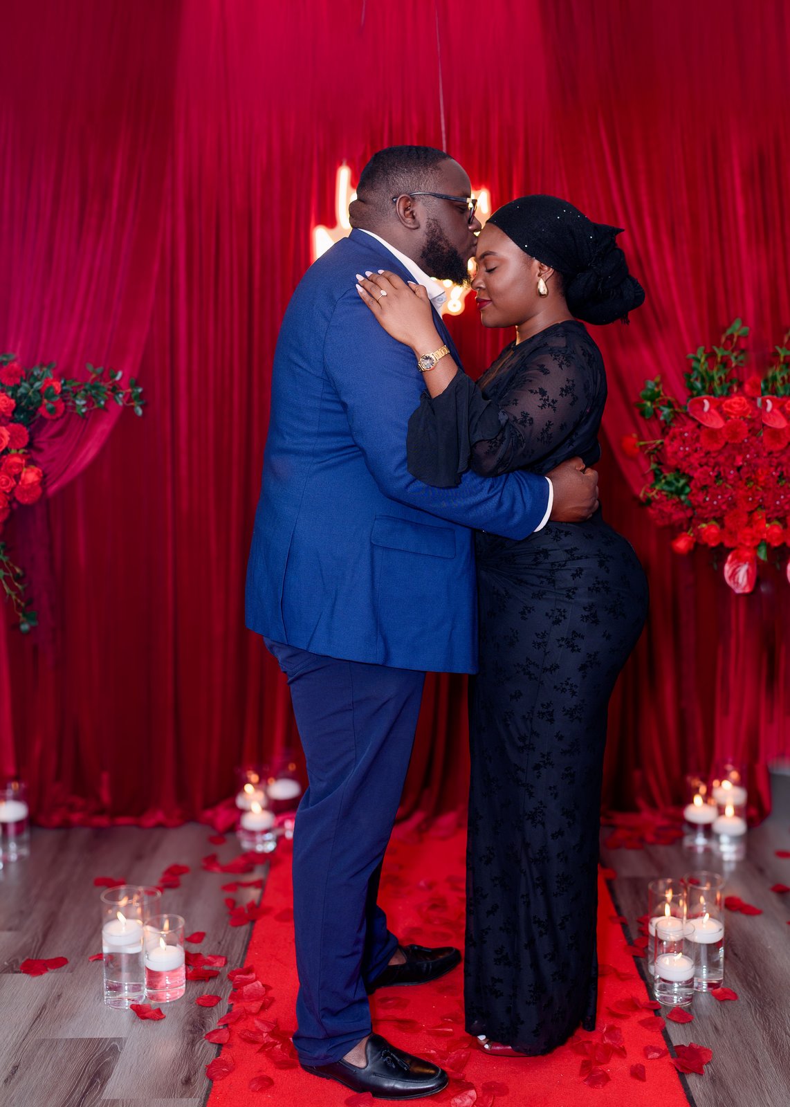 The couple embracing forehead-to-forehead in front of the red curtain backdrop with rose clusters and candles on the floor
