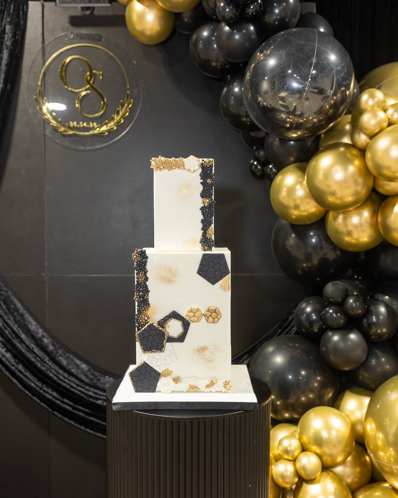 Two-tier black and gold geometric birthday cake against the balloon backdrop