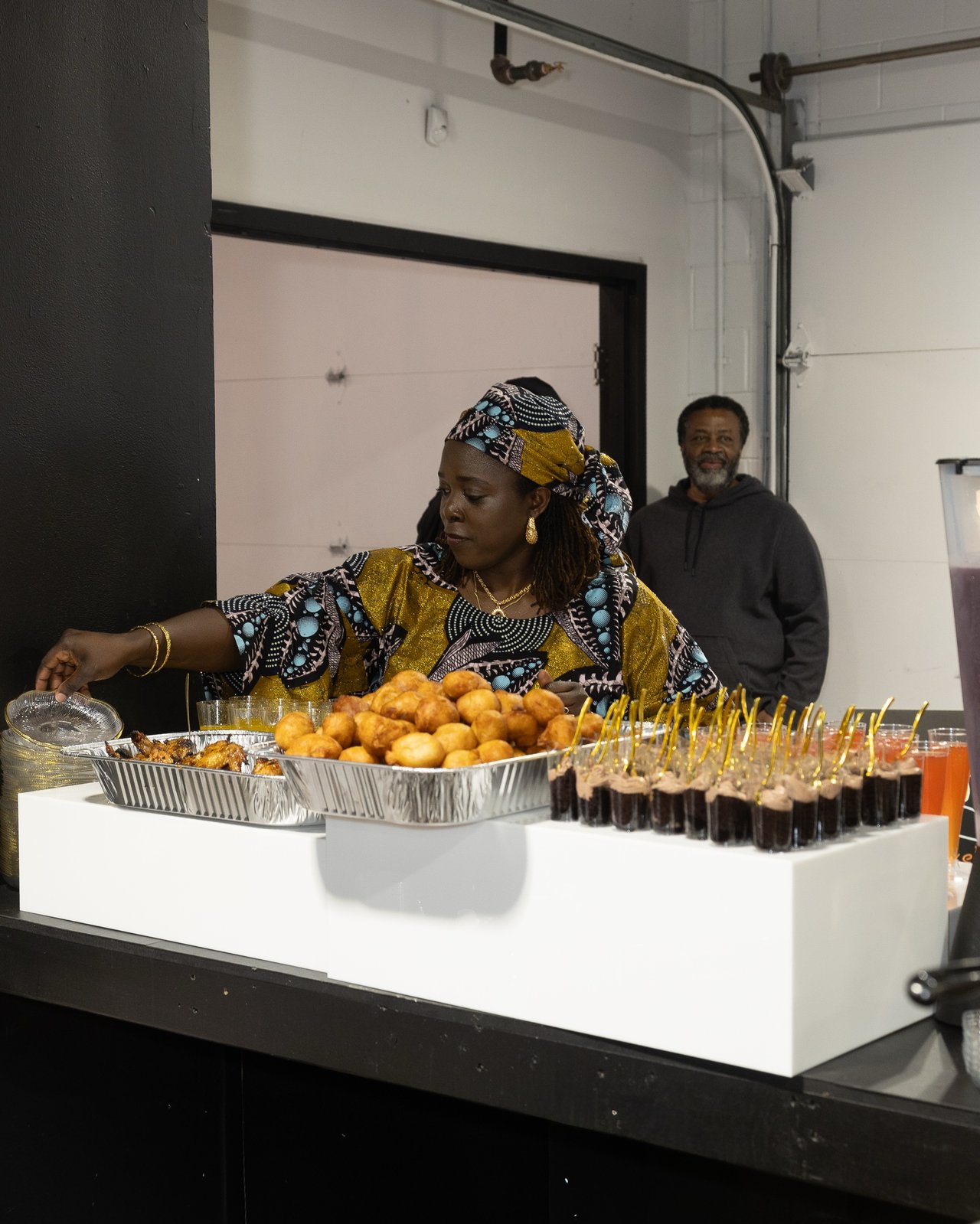 Guest in an Ankara headwrap reaching for a tray of puff-puff and dessert shooters