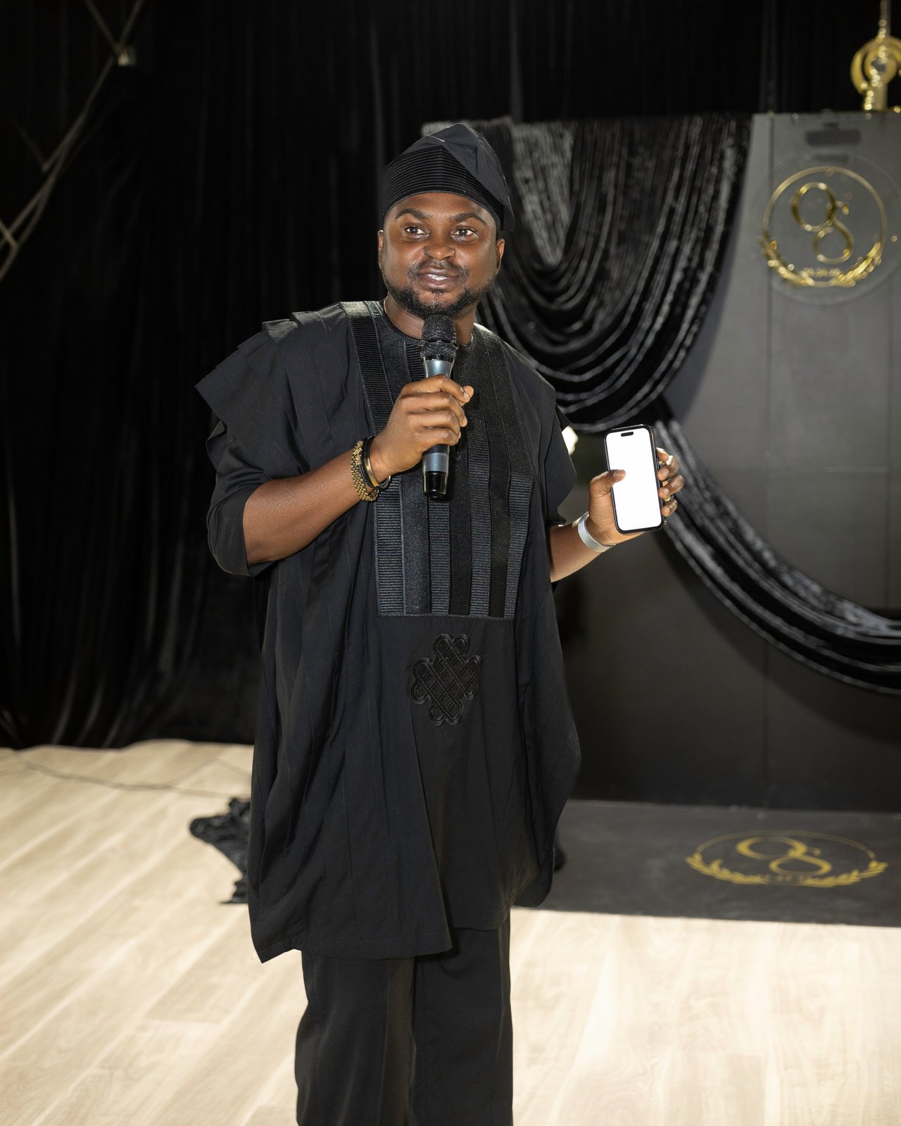 Host in a black embroidered agbada and cap speaking on the microphone in front of the OS monogram