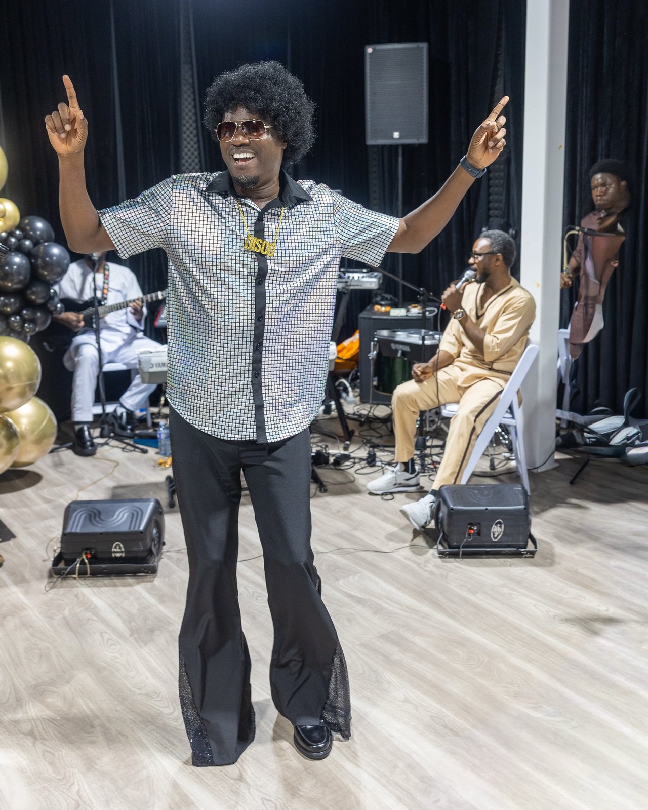 Guest in a disco shirt and afro wig dancing with arms raised in front of the live band