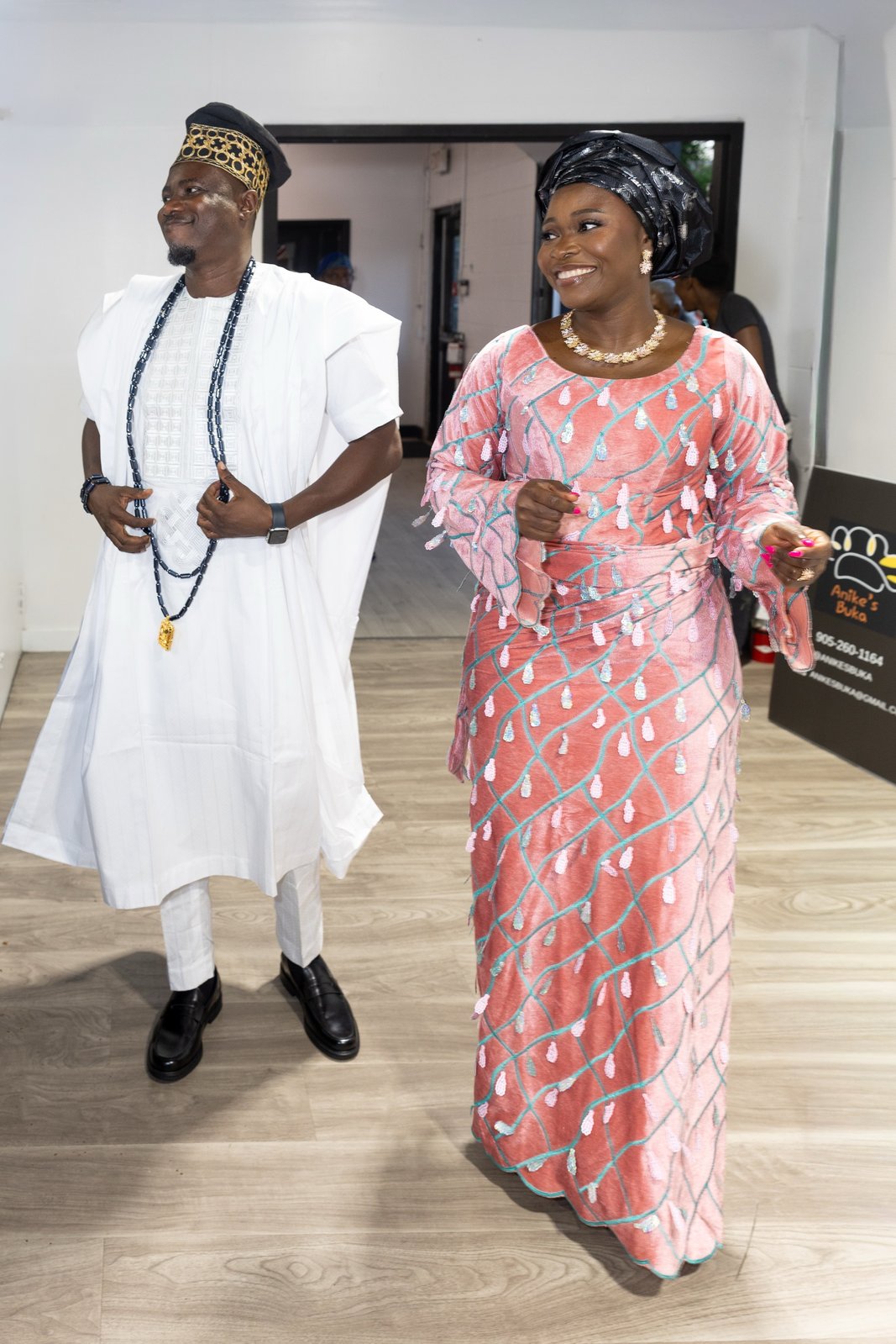 Couple in traditional Yoruba attire walking together, he in a beaded white agbada and cap, she in a coral beaded dress and gele