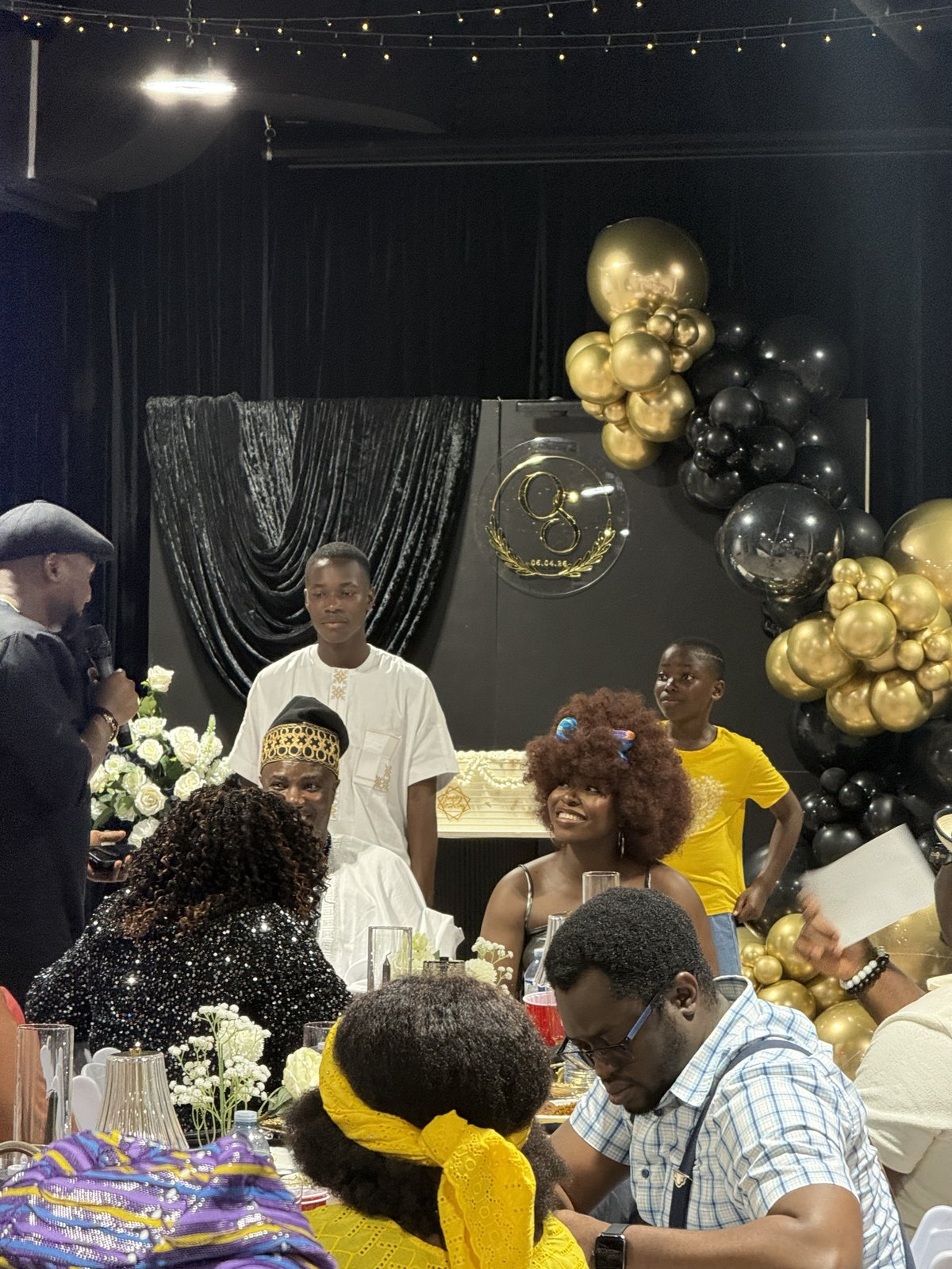 Wide view of guests gathered around the head table beneath the gold and black balloon arch