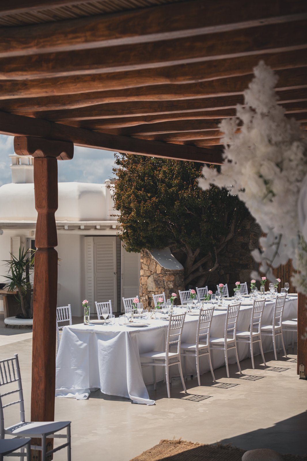 Reception table set under a wooden pergola with hanging white florals, overlooking the sea before guests arrive