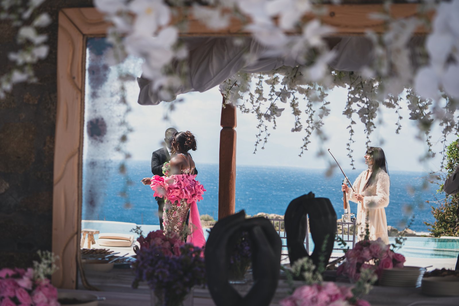 The couple embracing during the ceremony, framed by hanging white florals with a violinist and the sea beyond