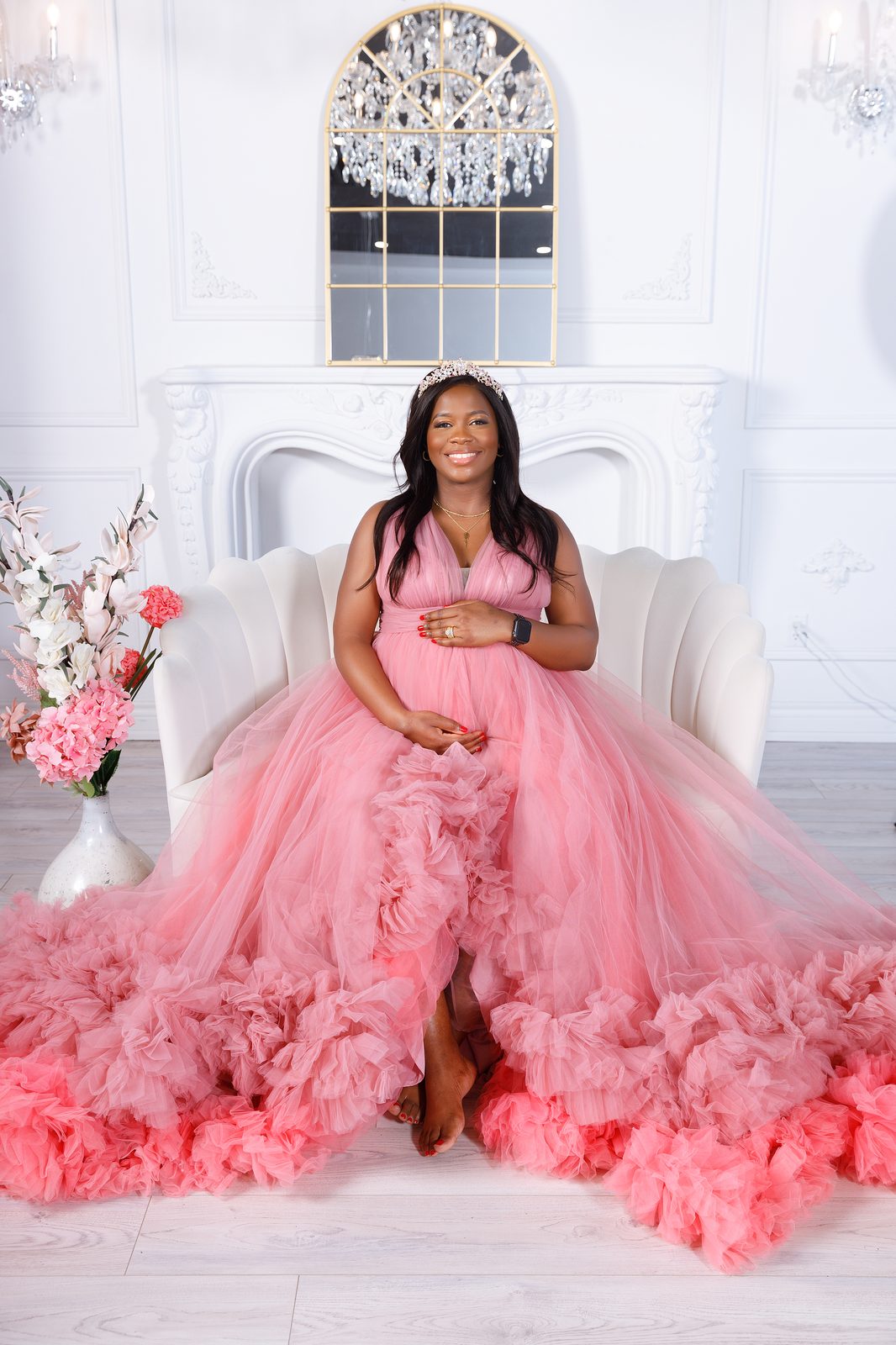 Expecting mother in a blush tulle gown and tiara beside a crystal chandelier